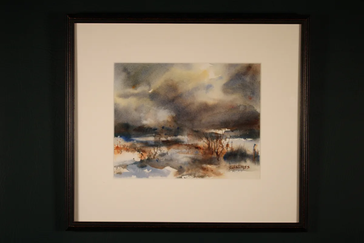 Framed watercolor winter landscape in blue-gray, brown, rust, and white tones. Snow-covered ground stretches beneath a dramatic cloudy sky, with sparse dark vegetation and distant landscape forms breaking through the wintry scene.